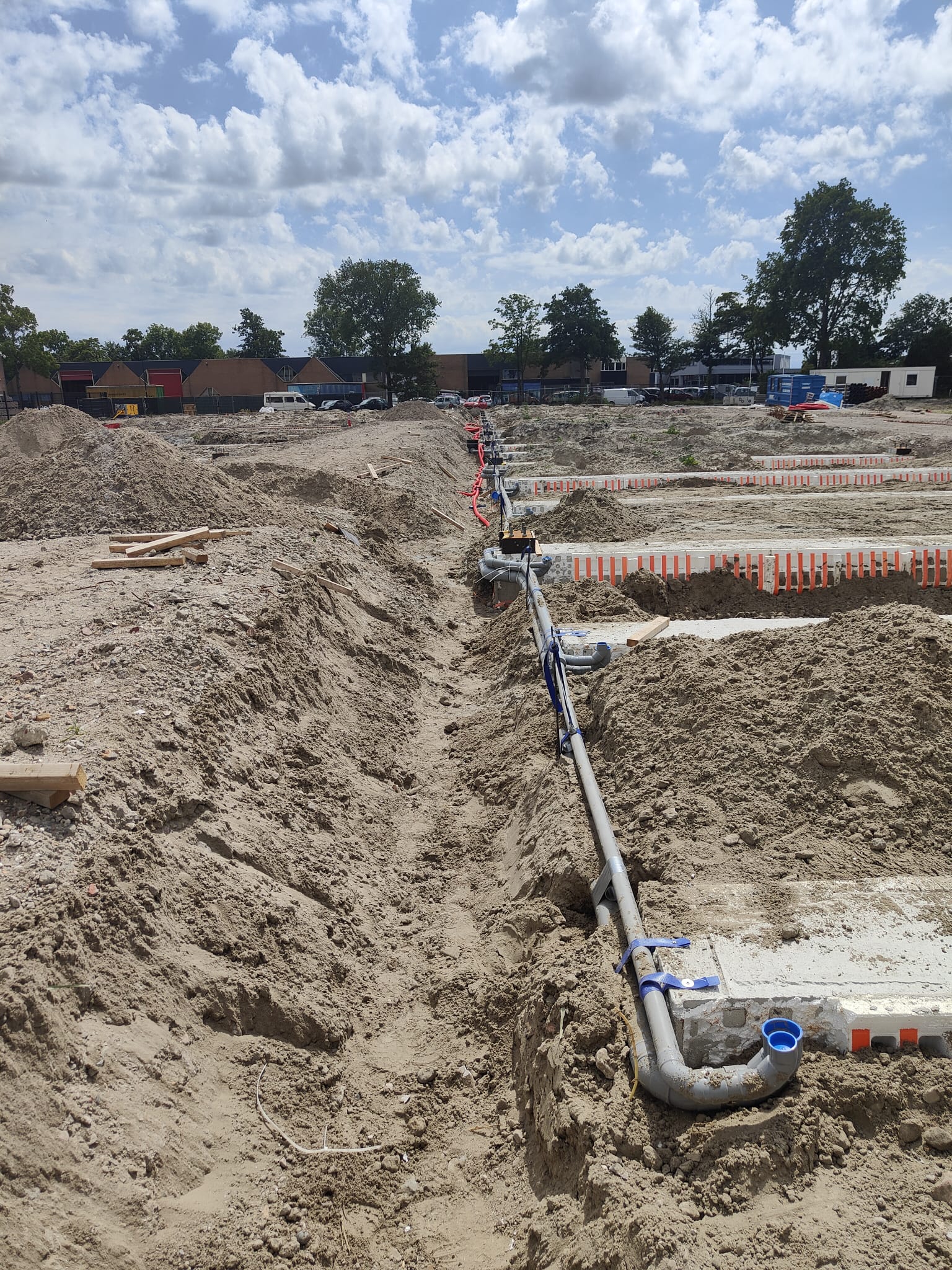 Een sleuf op een bouwterrein met een grijze rioolbuis en hulpstukken, onder een blauwe lucht
