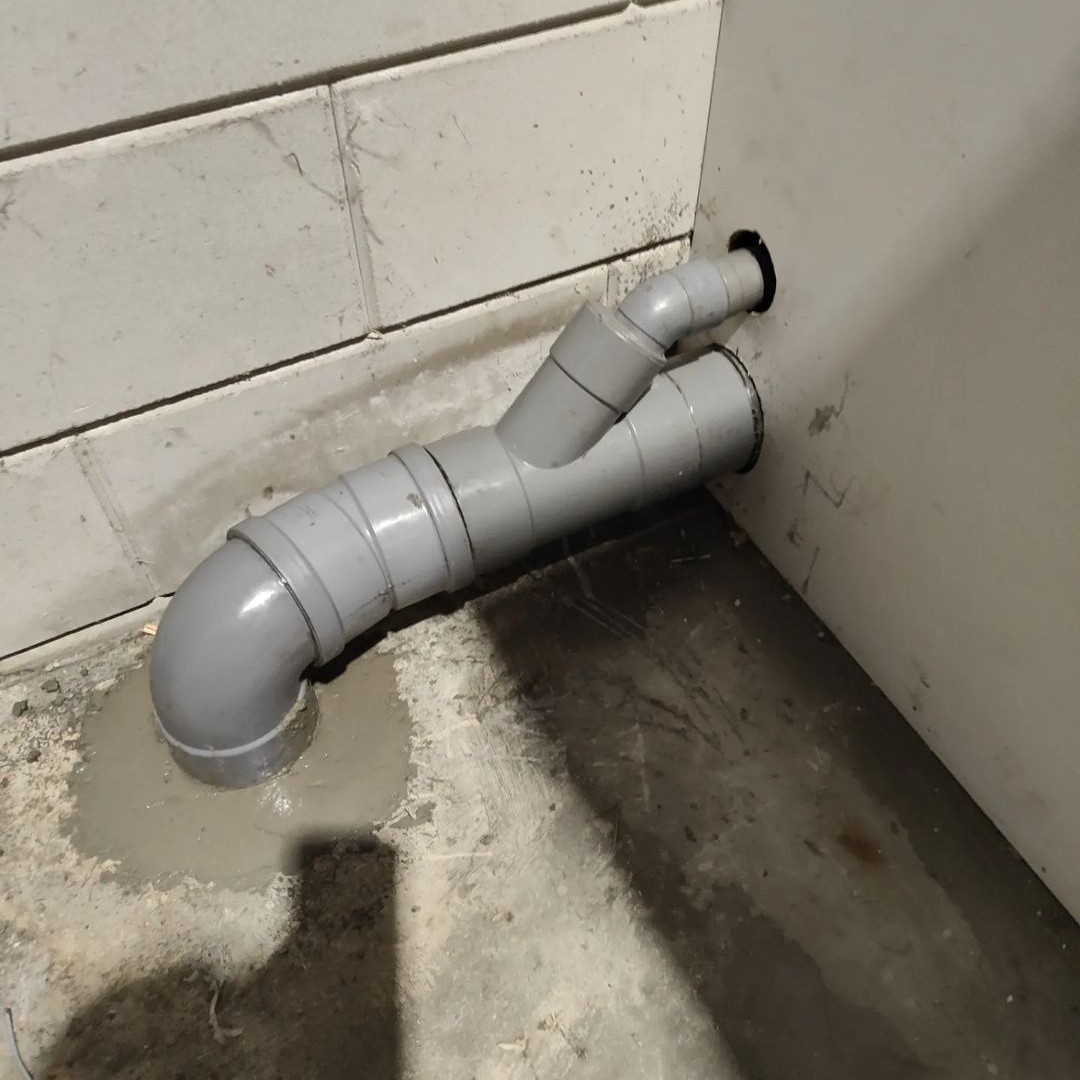 Grijze pvc-afvoer met bochten en een Y-stuk uit een betegelde wand op vloerhoogte