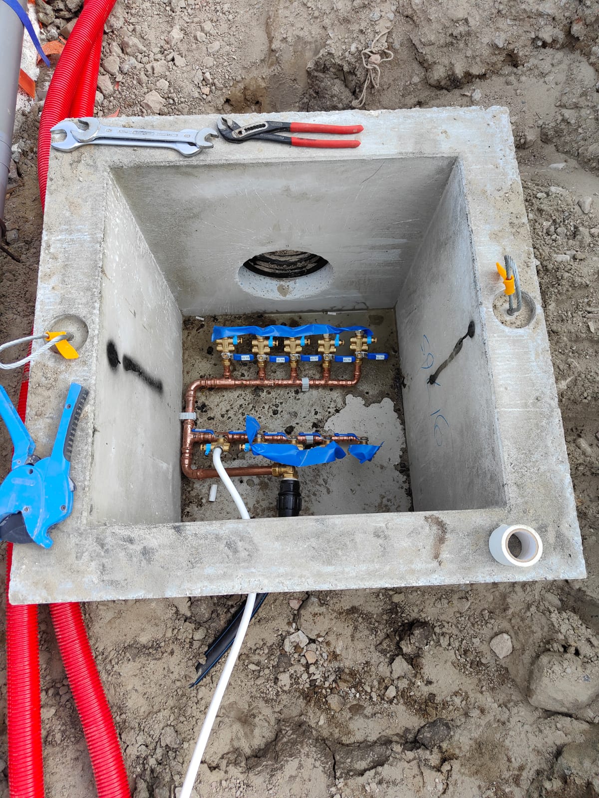 Koperen waterverdeler met blauwe afsluiters in een betonnen put in de grond, gereedschap op de rand