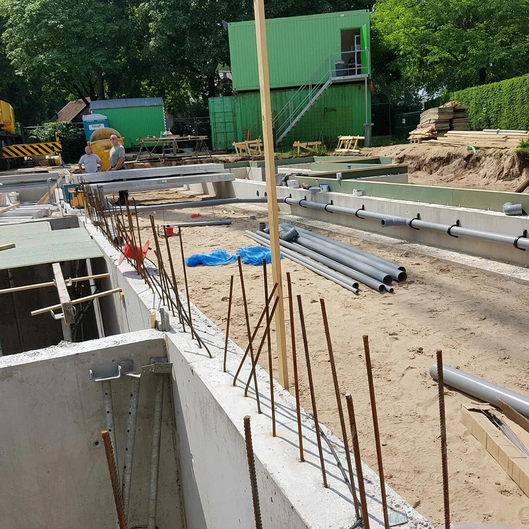 Bouwterrein met betonnen funderingswanden, grijze buizen en bouwketen op de achtergrond
